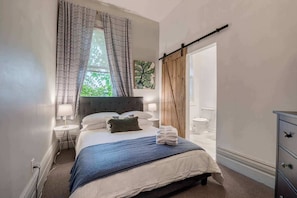 A bedroom with a bed, bedside lamps, a window with curtains, a wooden door, and a dresser. - Casa Sherbourne Hotel - Modern Industrial Suite (Toronto)