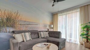 Ein modernes Wohnzimmer mit einem grauen Ecksofa, einem runden Couchtisch, einer Lampe und einem großen Fenster mit durchsichtigen Vorhängen.