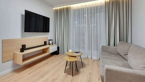 A modern living room with a sofa, a TV mounted on the wall, a wooden console table, and a small round table with a book. - Grano Residence 1 | Riverside | Parking (Gdańsk)