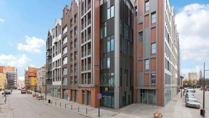 A modern multi-story building with a glass facade, a pedestrian walkway, and a parking area. - Grano Residence 1 | Riverside | Parking (Gdańsk)
