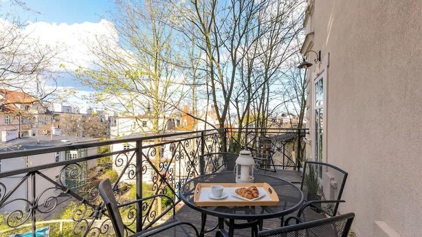 Een balkon met een tafel, stoelen, een dienblad met een croissant en een kopje, en uitzicht op gebouwen en bomen.