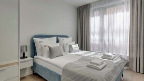 Quarto de hotel moderno com cama grande, roupa de cama branca, cabeceira azul e vista para a cidade através de cortinas leves.