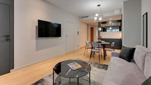 Ett modernt vardagsrum med en platt-TV, ett runt soffbord i glas, en soffa och en matplats med stolar.