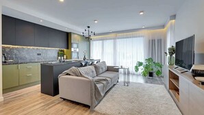 A modern kitchen with a central island, a sofa, and a TV in a living area with large windows and a plant. - Aura Island 90 Deluxe | River View | Parking (Gdańsk)