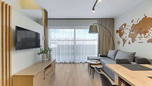 Ein modernes Hotelzimmer mit einem großen Flachbildfernseher, einem Holztisch, einem grauen Sofa, einem Esstisch und einer Weltkarte an der Wand.