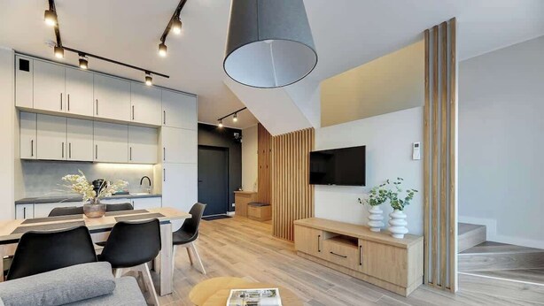 Eine moderne Küche mit weißen Schränken, ein Essbereich mit Tisch und Stühlen, ein Wohnzimmer mit Sofa und Couchtisch sowie ein an der Wand befestigter Fernseher.