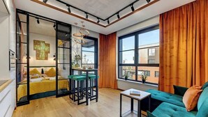 A modern living room with a sofa, a small table, a bar area with stools, a bed with a yellow headboard, and large windows with curtains. - Doki C80 | Gdańsk Shipyard & Foodhall (Gdańsk)