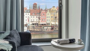 Une pièce avec vue sur une rivière, une chaise, une petite table avec une tasse et un journal, et une fenêtre avec des rideaux.