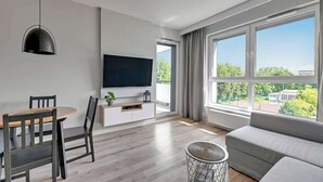 Een moderne woonkamer met een flatscreen-tv, een eettafel met stoelen, een bank en grote ramen met uitzicht op groen en gebouwen.