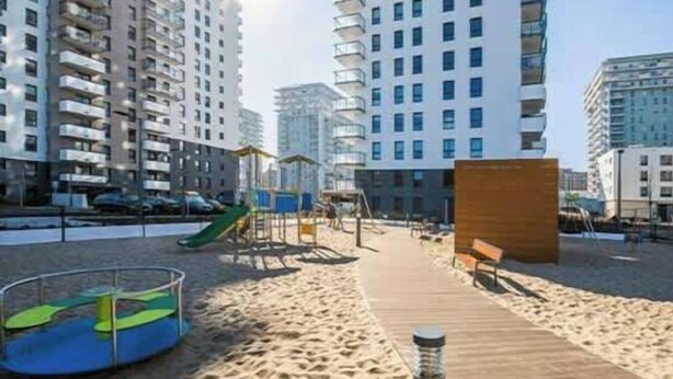 Een zandstrand met een houten looppad, speeltoestellen en hoge appartementen op de achtergrond.