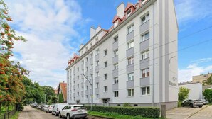 Un hôtel à plusieurs étages, avec une rangée de voitures garées, un trottoir pavé et un ciel dégagé.