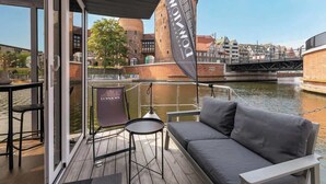 A deck with a sofa, chairs, and a table, overlooking a canal with buildings in the background.