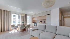 Ein modernes Wohnzimmer mit Sofa, Esstisch, Stühlen, einer Küchenzeile und großen Fenstern mit Vorhängen.