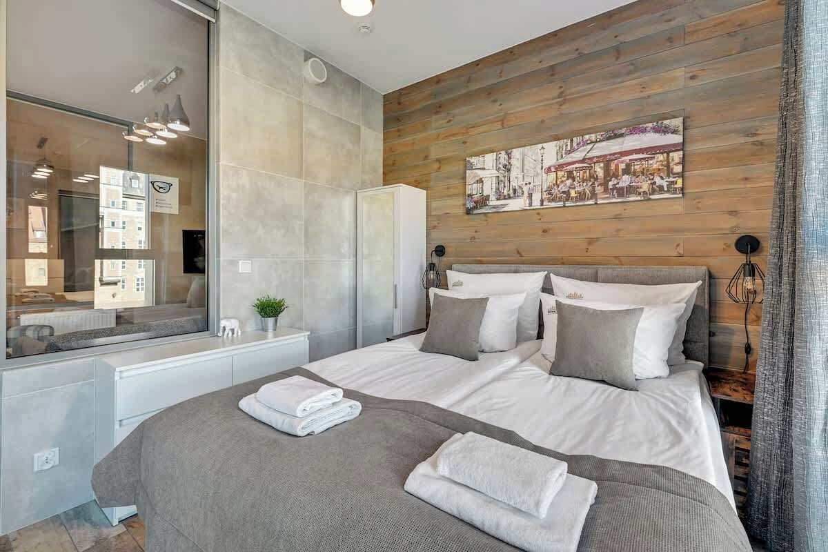 A modern bedroom with a large bed, a wooden accent wall, a mirror, and a view of the city.