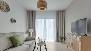 En moderne stue med sofa, sofabord, fjernsyn på et træskab og et stort vindue med gardiner.