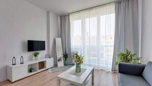Een moderne woonkamer met een flatscreen televisie, een witte salontafel, een grijze bank en grote ramen met dunne gordijnen.