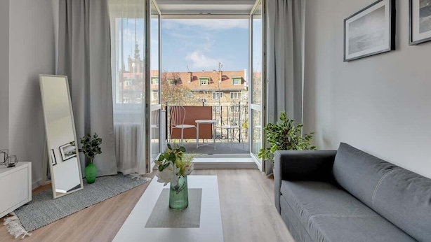 Een moderne woonkamer met een grijze bank, een witte salontafel, een groene vaas met bloemen en een groot raam met uitzicht op gebouwen en een balkon.