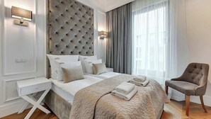 Una habitación de hotel moderna con cabecera capitonada, una cama grande con colcha color beige, una mesita de noche, una silla y un ventanal con cortinas.