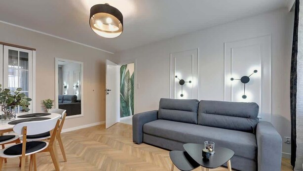 Ein modernes Wohnzimmer mit grauem Sofa, Holzstühlen, einem kleinen schwarzen Tisch und einer Deckenleuchte.