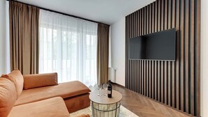 Een moderne woonkamer met een bank, een rond bijzettafeltje, een flatscreen televisie bevestigd aan een houten wandpaneel, en een raam met dunne gordijnen.