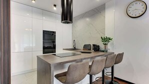 Een moderne keuken met witte kasten, een centraal eiland met barkrukken, een klok aan de muur en een marmeren achterwand.