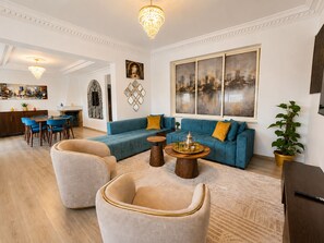 Ein modernes Wohnzimmer mit einer Eckcouch, einem Essbereich mit blauen Stühlen, einer Chandelieber und Kunstwerken an den Wänden.