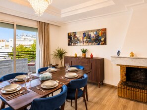 Ein Essbereich mit Holztisch, blauen Stühlen, einem Stadtbildgemälde, einem Kamin und einem Balkon mit Blick.