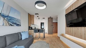 Et moderne opholdsrum med en sofa, et rundt sofabord, et fjernsyn monteret på væggen og et køkken i baggrunden.