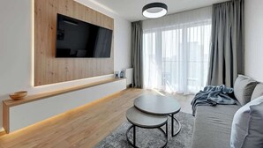 En moderne stue med et fladskærms-tv monteret på en træpanel, et rundt sofabord, en grå sofa og et stort vindue med lette gardiner.