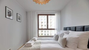 Hotelkamer met een bed, witte gordijnen, een raam met uitzicht op gebouwen en ingelijste foto's aan de muur.