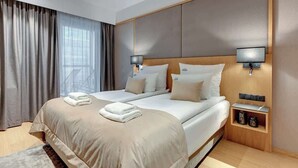 Un lit double avec une tête de lit en bois, deux tables de chevet avec des lampes, une grande fenêtre avec des rideaux et une petite étagère en bois sur laquelle sont posés divers objets.