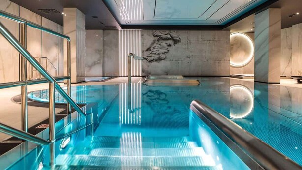 Une piscine intérieure moderne, dotée d’une paroi en verre, d’un mur en marbre orné d  ’une sculpture en relief et d’un luminaire circulaire.