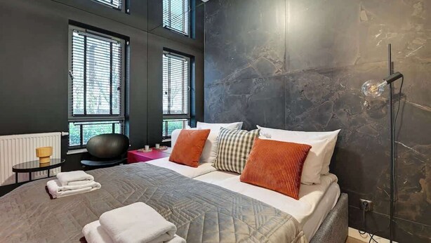 Una habitación de hotel moderna con una cama grande, varios almohadones, una lámpara de pie y un sillón de masajes.