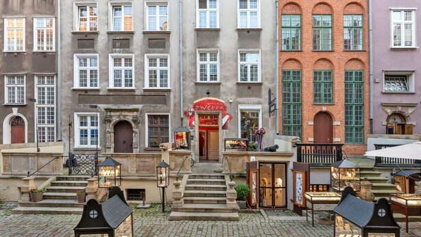 Ein historisches Gebäude mit einem zentralen Eingang und einer Treppe, die hinaufführt, sowie ein Geschäft mit rotem Markisenvorhang.