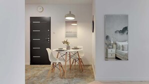 Una habitación moderna con zona de comedor, una puerta negra, un reloj montado en la pared, un cuadro enmarcado y una cama con ropa blanca.