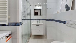 Un baño moderno con cabina de ducha de vidrio, azulejos blancos, un lavabo con espejo y un inodoro.