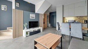 Ein modernes Wohnzimmer mit Flachbildfernseher, Couchtisch aus Holz, grauen Sesseln und einem Essbereich mit Küche im Hintergrund.