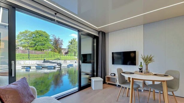 A modern living room with a large sliding glass door, a flat-screen TV mounted on the wall, a dining table with chairs, and a cozy seating area with a sofa and cushions.