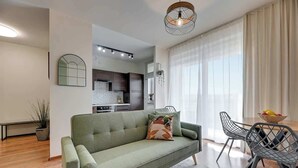 Ett modernt vardagsrum med en soffa, en matplats med stolar, ett kök i bakgrunden och ett stort fönster med gardiner.
