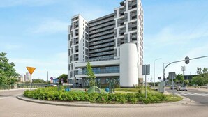 A modern multi-story building with a central cylindrical structure, surrounded by a landscaped area with bushes and a playground. - Gdynia Modern Tower | Great City View | 13 floor (Gdynia)