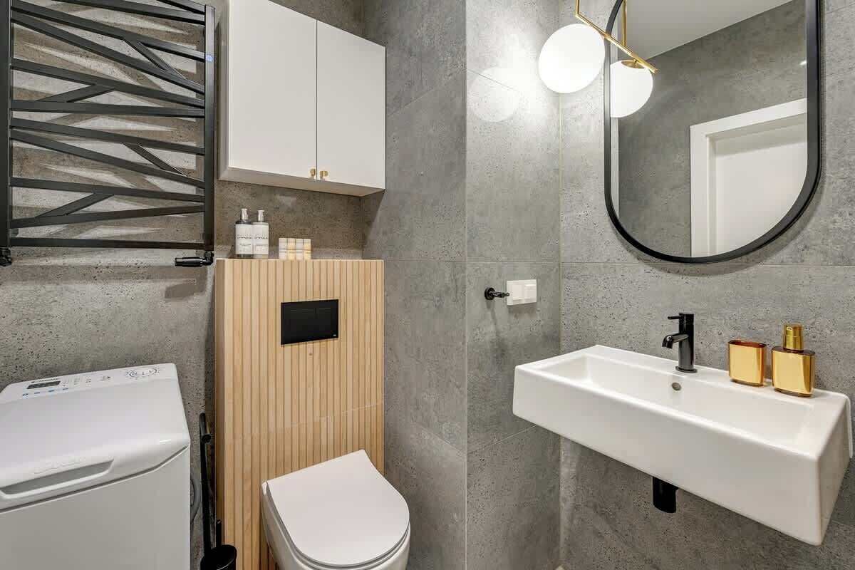 Un baño moderno con lavadora integrada, inodoro, mueble de madera, lavamanos con detalles dorados y un espejo grande.