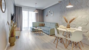 A modern living room with a sofa, dining table, and chairs, a potted plant, and a geometric wall design. - Aura Island 66 | Riverside | City Center & Parking (Gdańsk)