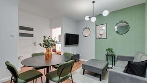 A modern living room with a round dining table, green upholstered chairs, a grey sofa, and a TV mounted on the wall. - Aura 196B | Riverside | City Center (Gdańsk)