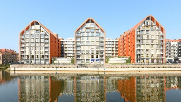 A modern building with a unique triangular roof design, located near a body of water. - Aura 196B | Riverside | City Center (Gdańsk)