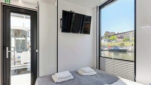 Moderni hotellihuone, jossa on sänky, seinälle asennettu televisio, parveke, jolta on näkymä kaupunkiin, sekä ovi, joka johtaa parvekkeelle.