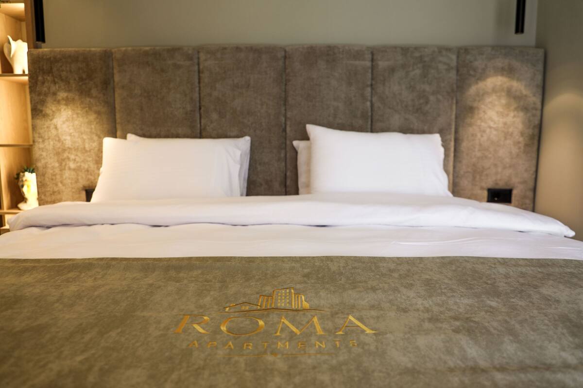 A neatly made bed with a headboard, a logo for 'ROMA APARTMENTS' on the bedspread, and a nightstand with a lamp to the left.