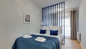 Un dormitorio con cama, almohadas, una colcha, un cuadro en la pared y una ventana con cortinas.