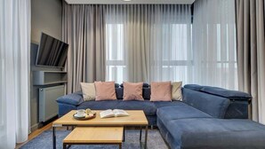 Un salon moderne avec un canapé d’angle, une table basse, un téléviseur à écran plat fixé au mur et de grandes fenêtres ornées de rideaux légers.