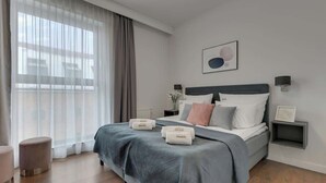 Une chambre à coucher moderne, avec un lit, des tables de chevet, une commode, une fenêtre avec des rideaux et une œuvre d’art accrochée au mur.