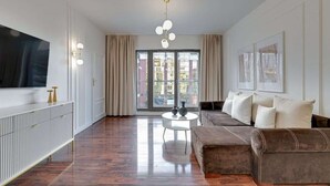 A modern living room with a sofa, a TV, a round coffee table, and a large window with curtains. - Waterlane E6 | Spa, Pool & Gym | River View (Gdańsk)